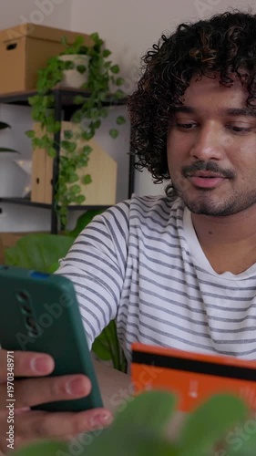 Vertical HD video of young adult Indian man paying online with credit card using mobile phone at home office. Freelancer managing digital transaction during remote work. Online shopping concept.