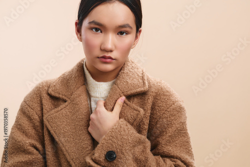 Tableau sur toile Asian woman wears a warm beige coat against a neutral studio backdrop, creating a timeless portrait that emphasizes calm confidence and minimalist fashion