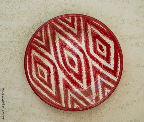 Traditional uzbek ceramic plate lagan with red atlas pattern hanging on stone wall. Rishtan Ceramics