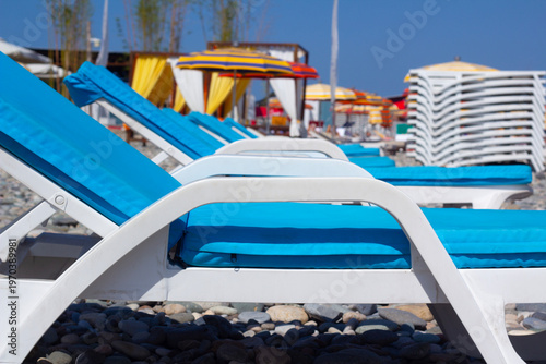 Horizontal defocused photo. Row of blue chaise longue, sun lounger, deck chair, colorful umbrellas, parasols on pebble beach of Batumi at Black sea coast. Selective focus. Tourism, relax, calm, travel