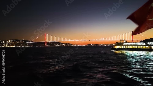 Cinematic Night View of Bosphorus Bridge in Istanbul with Waving Turkish Flag and Passenger Ferry Crossing the Strait, Beautiful City Lights Reflecting on Sea Water During Blue Hour and Twilight