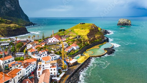 Aerial view of Porto da Cruz, Madeira, showcasing its unique black sand beach, natural volcanic pools, and charming coastal village against dramatic cliffs and the Atlantic Ocean.