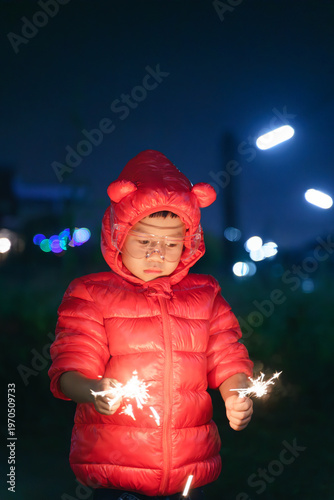 Child in a red hooded jacket and safety glasses holding a sparkler at night, creating a warm and festive glow.