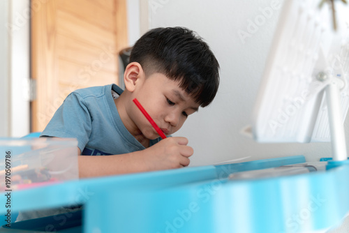 Close-up of an Asian boy focused on drawing or writing with a red pencil at a blue desk, symbolizing creativity, concentration, and learning.