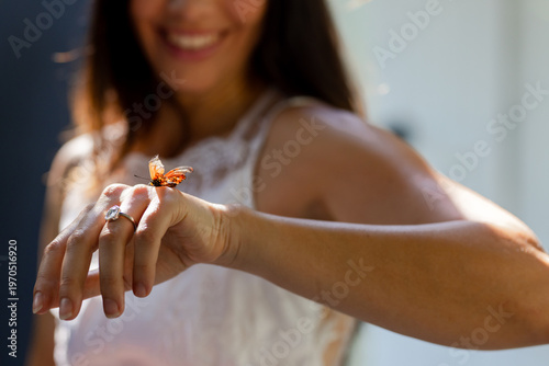 Adult woman holding left hand to camera in sunlit room white-lace with orange butterfly on ring