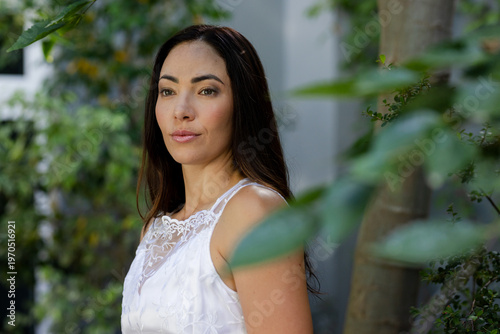 Adult female posing, looking left in garden wearing white lace dress amid foliage, tree trunk, wall