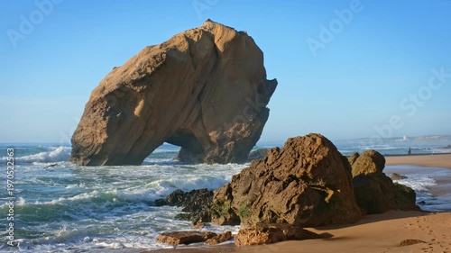 Penedo do Guincho is a large rock arch located at Praia da Santa Cruz in Portugal. The sight includes ocean waves crashing against sandy beach during sunset.
