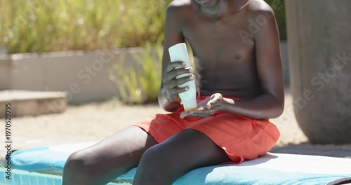 Sunny African American teen shirtless applying sunscreen into arms on towel protecting skin