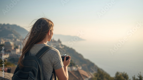 Young female traveler with backpack holding a camera and looking at a scenic coastal town view from a hilltop.