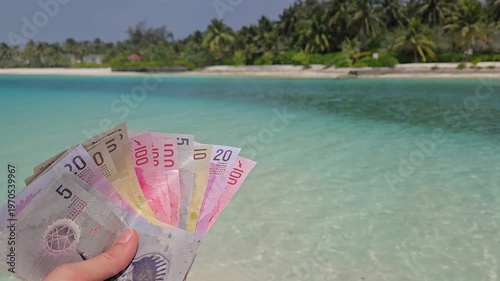 Stack of different denominations of Banknotes of Maldives, Maldivian rufiyaa in hand with maldivian island and ocean in background. Expensive travel concept