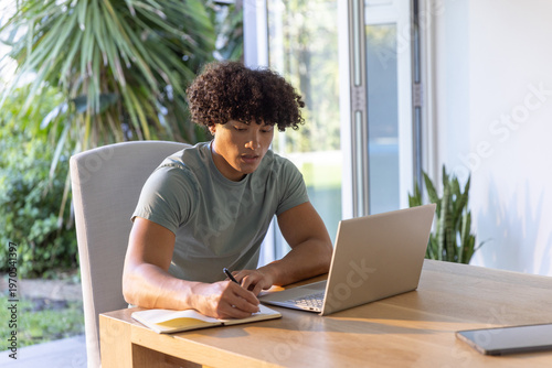 Foto African American male writing in notebook with pen beside open laptop by sliding