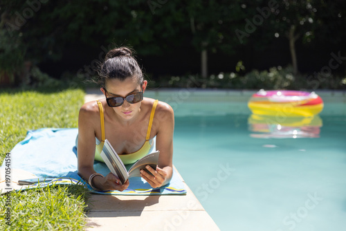 Woman lying on stomach at pool reading book with shades, yellow straps, bright ring nearby