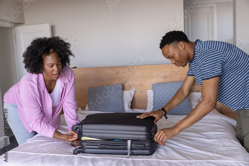 African American couple leaning over queen-size bed, closing black hard-shell suitcase together