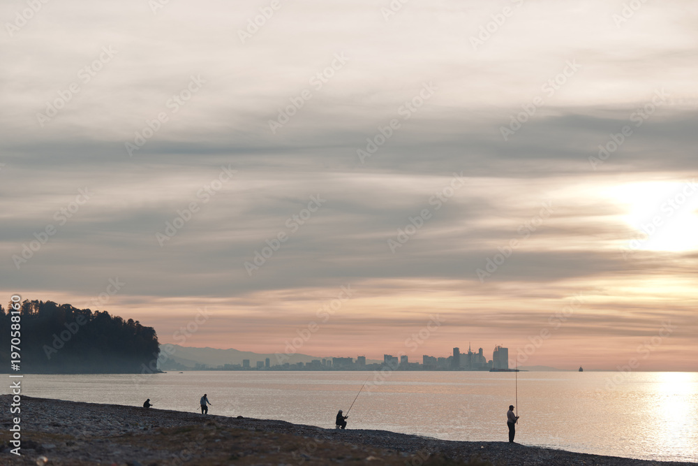 Obraz premium Beach sunset with silhouettes of fishermen along the shore, calm water and distant skyline, warm glow and golden light creating a tranquil coastal scene for travel and nature photography.