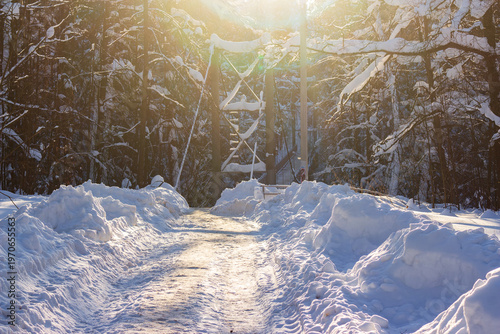 Sunny winter path winding through deep snow drifts towards a pedestrian bridge in a serene forest, bathed in golden light