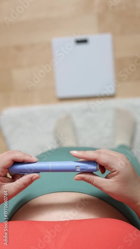 A vertical close up of a young woman screwing a needle on pre-filled pen injector with semaglutide to prepare it for taking medicine on floor and scales background