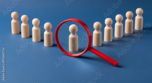 Recruitment and selection concept with a red magnifying glass focusing on one wooden peg figure among a group on a blue background