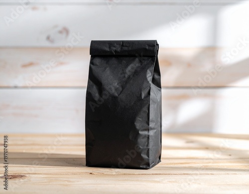 Minimalist black paper bag with a folded top, resting on a light wooden table against a white plank wall, ideal for eco-friendly packaging, food, coffee, or small gifts