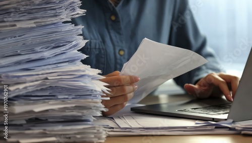 A person sorting through a large stack of paperwork while using a laptop, indicating a busy work or office environment.