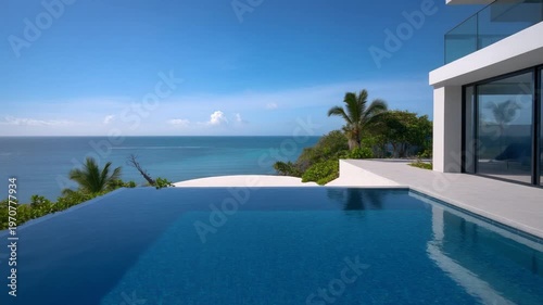 Modern luxury villa with a large infinity edge swimming pool overlooking a tropical turquoise ocean, featuring a minimalist white architectural design, palm trees, and clear blue sky.