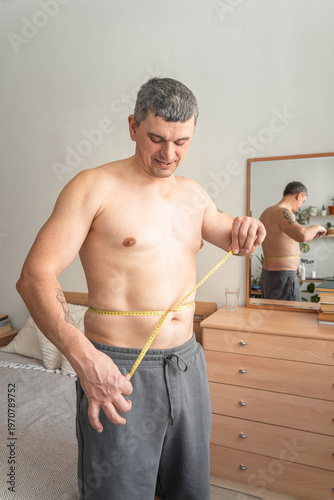 Man measuring his bare waist with a tape measure, monitoring progress for weight loss, diet, and fitness goals. Real person, self-acceptance, authentic lifestyle