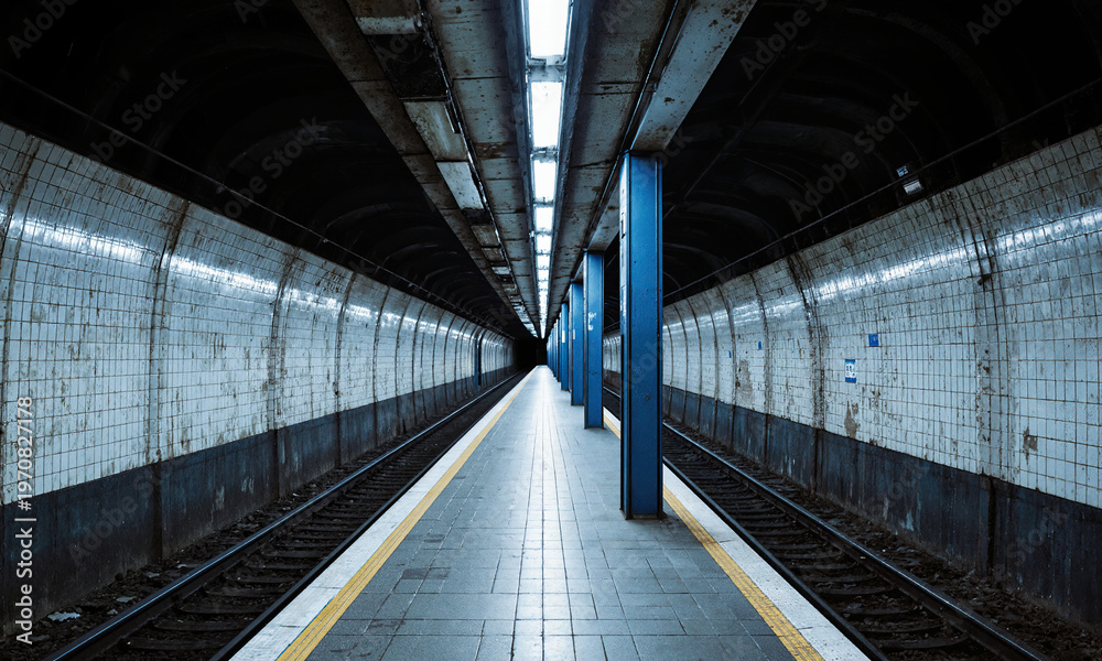 Fototapeta premium Subway station platform stretching into a dark tunnel with grimy tiled walls and blue pillars in a deserted underground terminal