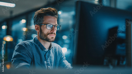 Man working on computer in low light office environment representing late work hours and technology driven tasks