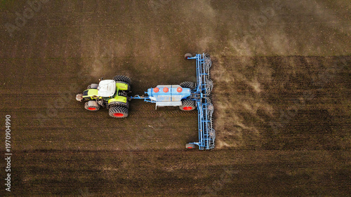 Aerial view of a green and white tractor with orange wheels plowing a brown field using a blue seed drill, showcasing agricultural machinery in action