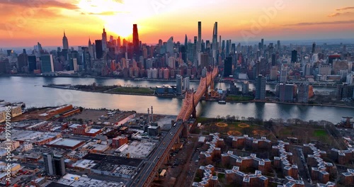 Approaching the Queensboro Bridge with cars heading to and from the structure. Aerial perspective on the stunning New York skyline against orange sky at sunset.