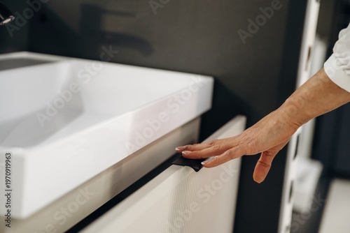 Close-up of a hand reaching for a modern bathroom cabinet drawer with a white sink and faucet in a contemporary interior design setting