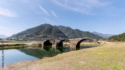 Time-lapse of the ancient three-arch stone bridge under blue skies with spring reflections and visible signs of age and resilience, Wuyuan County, Jiangxi Province, China