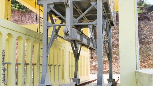 Side view shows hydraulic gate and lifting system on dam. Steel mechanism stands on tracks near concrete wall. Functional hydraulic gate belongs to lifting system. Industrial structure is ready.