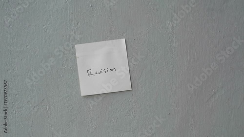 Hand sticking a white square paper note with the word Revision on a textured wall.