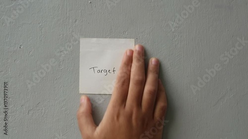 Close-up of Hand Sticking White Sticky Note with 'Target' Written on Gray Wall
