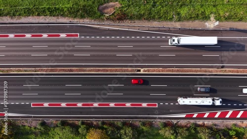 Overhead top-down view of a dual carriageway highway showing multiple lanes of traffic with red and white chevron road markings, white dashed lane lines, trucks and cars in motion