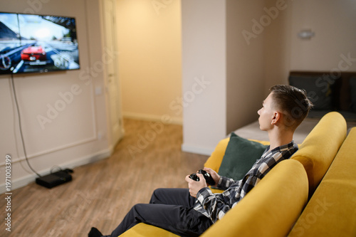Teenage Boy Playing Car Racing Video Game on Large TV Screen at Home, Young Gamer Using Controller on Yellow Sofa. Gaming Hobby, Leisure Concept, Modern Interior with Gaming Console, Virtual Reality