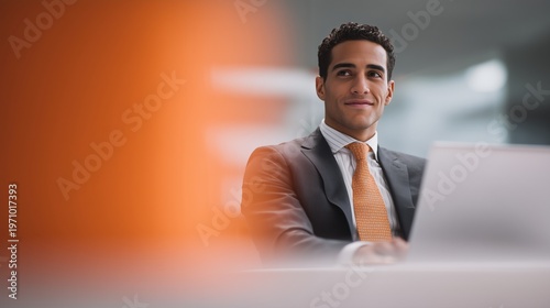 A young Hispanic man with dark hair sits at a desk, smiling while working on a laptop. An orange object is blurred in the foreground.