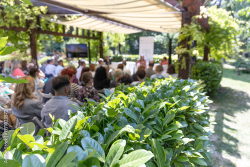 Outdoor business conference with professional audience attending presentation in green garden venue under canopy, sustainable corporate event setting