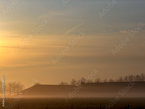 Misty sunrise over rural countryside with silhouetted trees and farmland in golden morning light