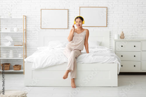 Beautiful young happy Asian woman in headphones listening music on bed at home