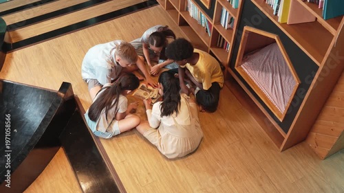 Group of diverse children sitting in circle while reading a book at library. Top view of girl sharing a magazine while pointing at interested topic and talking with multicultural friends. Edification