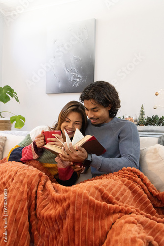 South Asian couple wearing striped sweater and blue pullover reading book on sofa, holding red mug