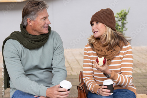 Middle-aged couple in scarves sitting on bench holding to-go cups and bitten apple by leather bag