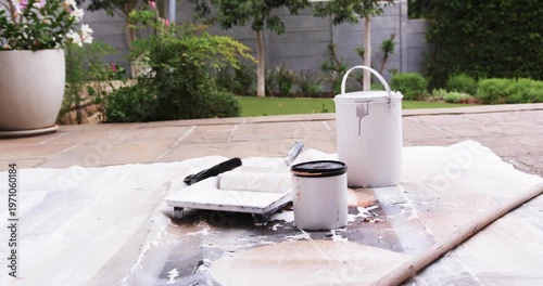 Camera pulling back revealing white bucket and roller, paint supplies on patio showing garden wall