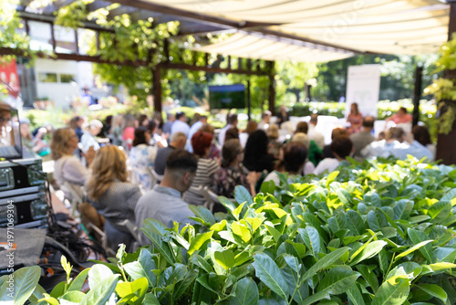 Outdoor business conference with professional audience attending presentation in green garden venue under canopy, sustainable corporate event setting