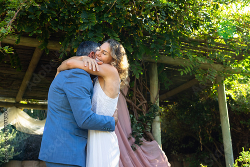 Couple embracing at garden pergola with pink drape, in white dress and blue suit, celebrating vows