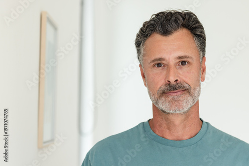 Middle-aged male standing by pale hallway wall wearing teal crew-neck T-shirt with framed art