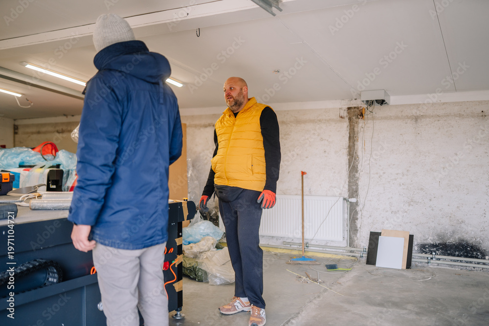 Fototapeta premium Two men discussing renovation plans inside a garage workspace, one wearing a yellow vest and the other in a blue jacket, with tools and materials visible in the background