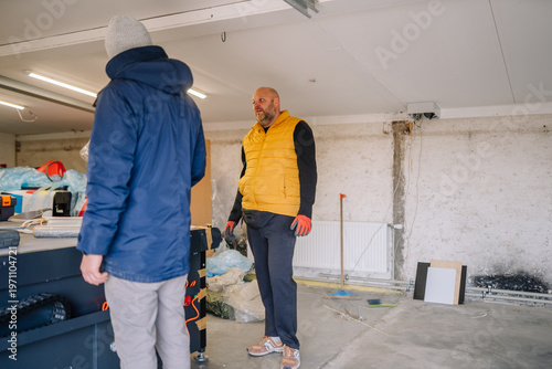 Two men discussing renovation plans inside a garage workspace, one wearing a yellow vest and the other in a blue jacket, with tools and materials visible in the background