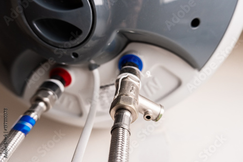 Close-up of a water heater's connection points. Red and blue pipes are visible, indicating hot and cold water lines. The heater has a sleek, modern design.